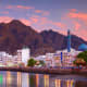 Muscat waterfront at sunset with white buildings, blue-domed mosque, minaret, and dramatic rocky mountains reflected in calm 